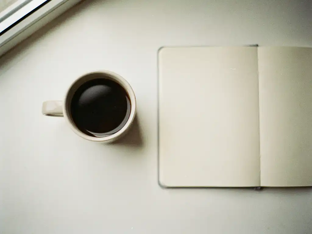Keramische kop zwarte koffie naast een open notitieboekje op een wit bureau, fotografeerd van bovenaf in zacht natuurlijk licht.